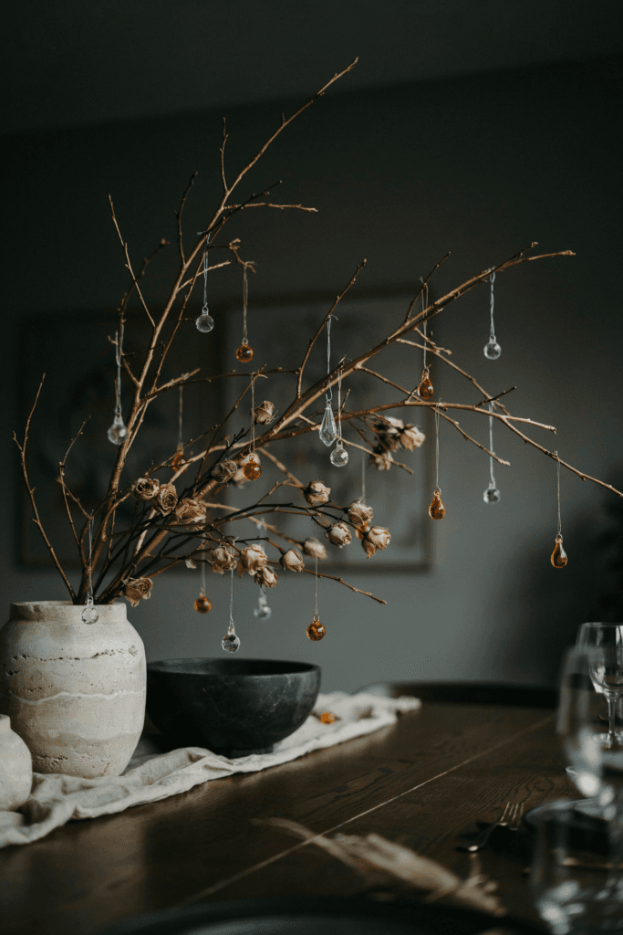 Tall branches spray-painted in matte gold and arranged in heavy stone vessels, with tiny dried rosebuds, crystal drops, and amber glass ornaments hung from the branches like a living mobile above the table