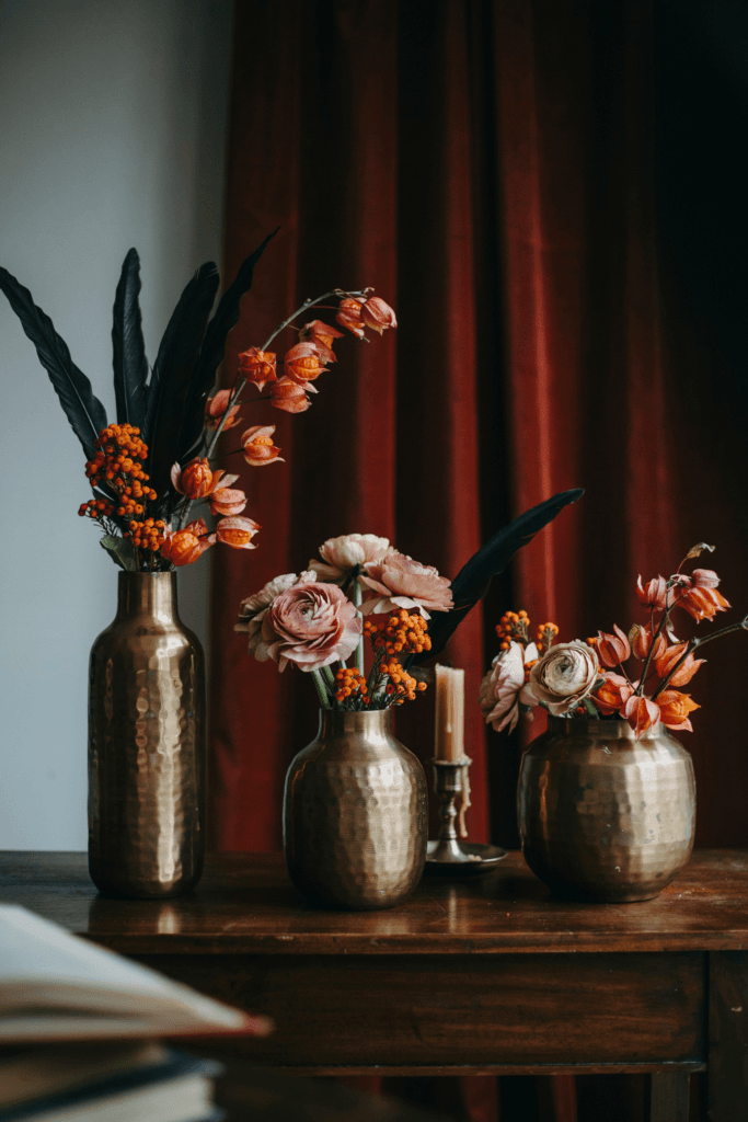 Whimsical arrangements combining dark feathers, orange hypericum berries, orange bittersweet branches, and dusty rose ranunculus in hammered bronze vases