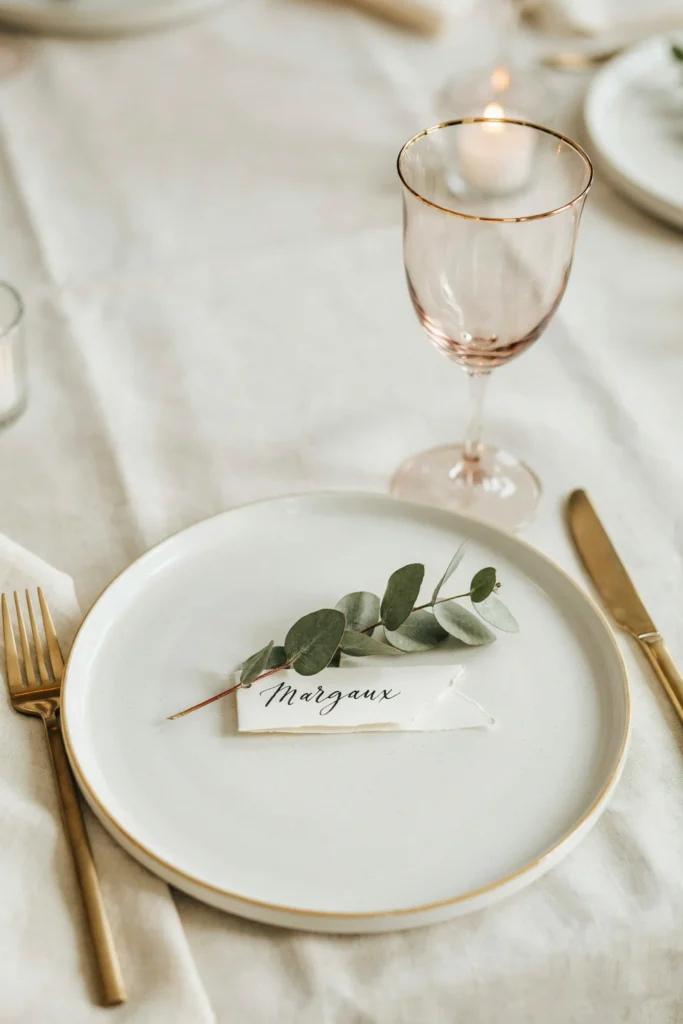 A close-up of an elegant place setting featuring a blush-tinted goblet on a white charger plate with gold flatware and a sprig of greenery as a place card holder