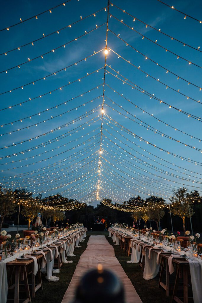 An outdoor aisle photographed looking up, with hundreds of warm white string lights forming a dense canopy overhead against a deep blue dusk sky