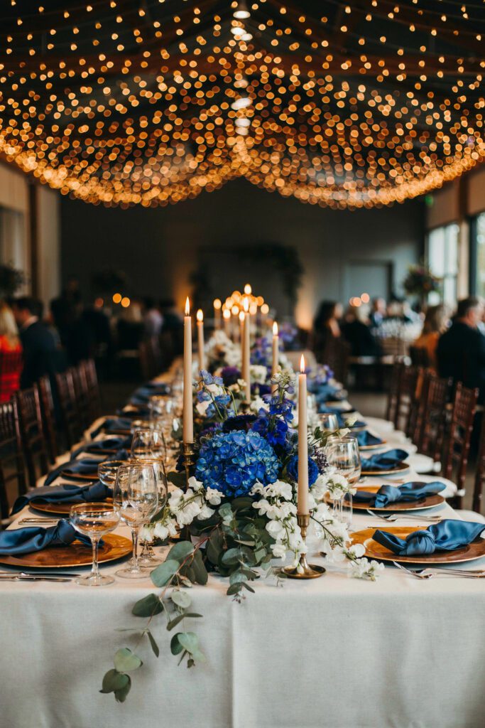 A reception table featuring a deep royal blue floral centerpiece with white sweet peas and trailing eucalyptus, photographed against a warm string-light background, with blue silk napkins and gold charger plates