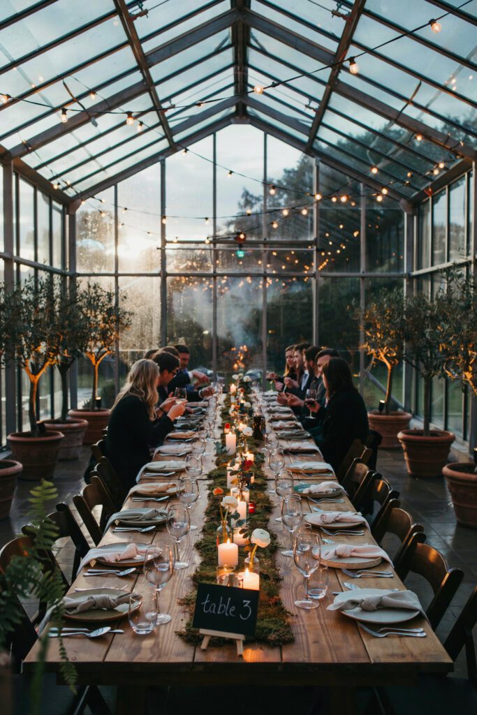 An indoor reception in a glass greenhouse venue, with potted ferns and small trees lining the perimeter, string lights overhead, and long wooden dining tables down the center with moss runners and candles