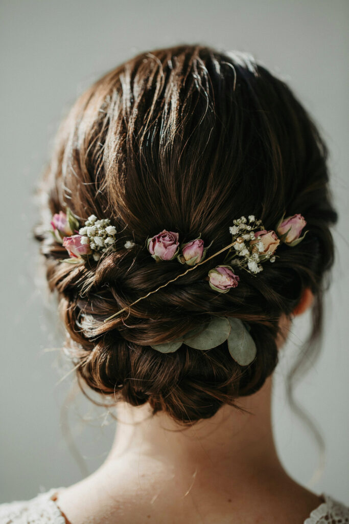 A close-up of a bridal updo with small dried flower pins scattered throughout the twist — tiny roses, baby's breath clusters, and a pressed leaf or two visible against dark hair in afternoon light