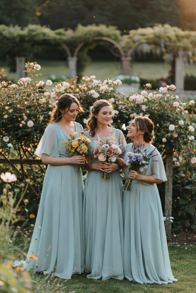 Three bridesmaids in flowing sage green chiffon dresses standing in a garden, each holding a loose wildflower bouquet in mismatched colors — one in soft yellow, one in blush, one in lavender — photographed in golden hour light