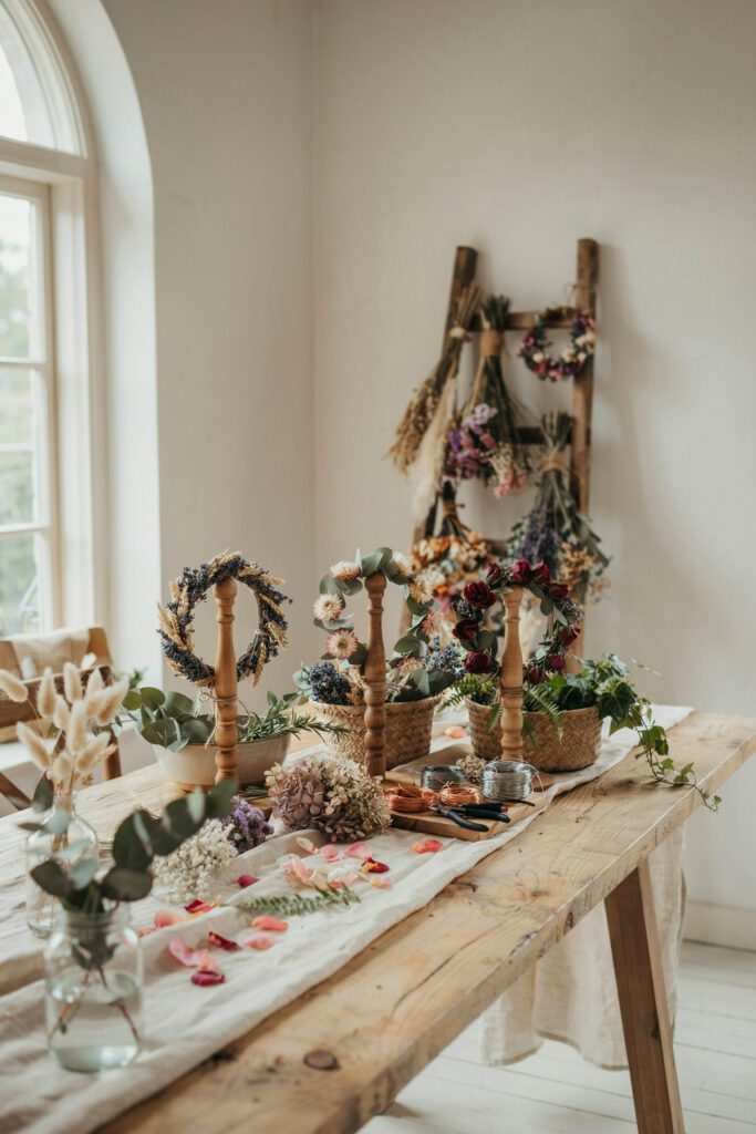  A long table set up as a floral crown station with small bundles of dried flowers, greenery, and wire in baskets, with three completed floral crowns displayed on small wooden stands