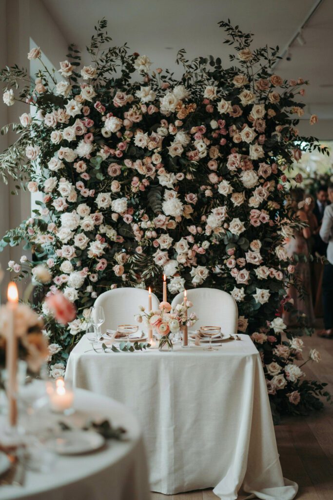 A sweetheart table for two positioned in front of a dense floral wall installation in ivory, blush, and sage, with the table itself set simply with white linens, tapered candles, and a small personal floral arrangement