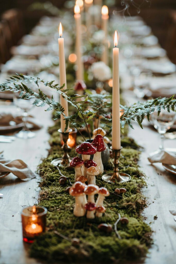 A reception table detail shot showing small ceramic mushroom and toadstool figures tucked into a moss table runner between candles and fern fronds, photographed at eye level so they look like they're growing from the greenery