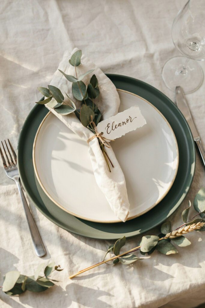  A place setting featuring a dark green charger plate with a cream dinner plate centered on it, a linen napkin folded into a leaf shape and tied with a sprig of eucalyptus, and a small handwritten place card tucked into the fold