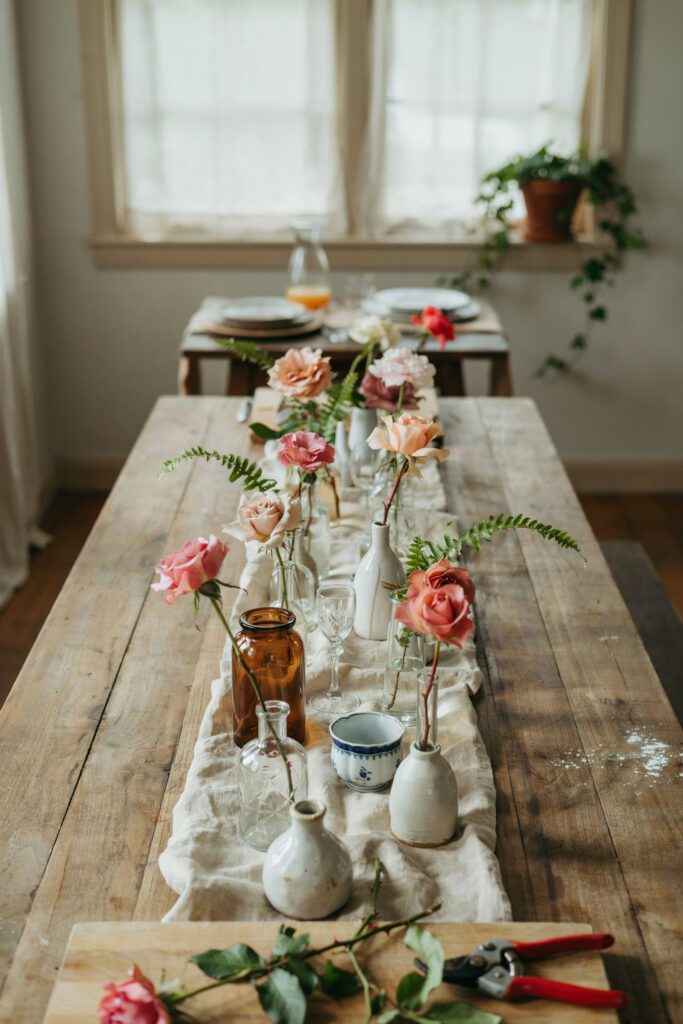 A farm table with a dozen small bud vases in varying heights clustered along the center, each holding one or two garden roses in blush, cream, and soft coral, with small fern fronds tucked between the vases