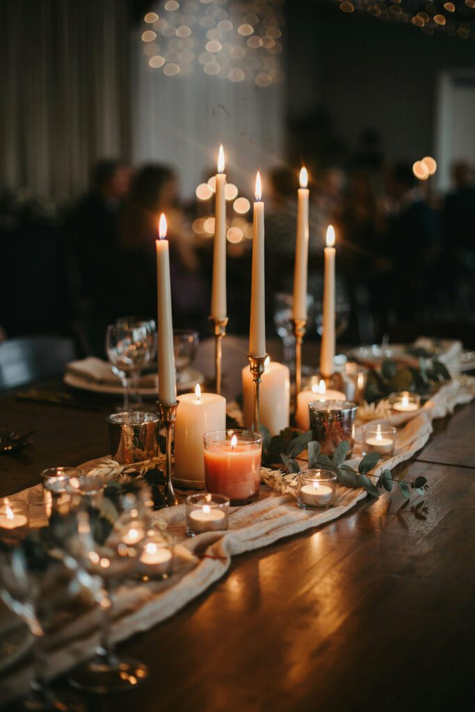 A close-up of a reception table with an arrangement of candle holders at four different heights — tall tapered candles in brass holders, medium pillar candles, short votives, and tea lights in glass — all lit, creating layered warm glow