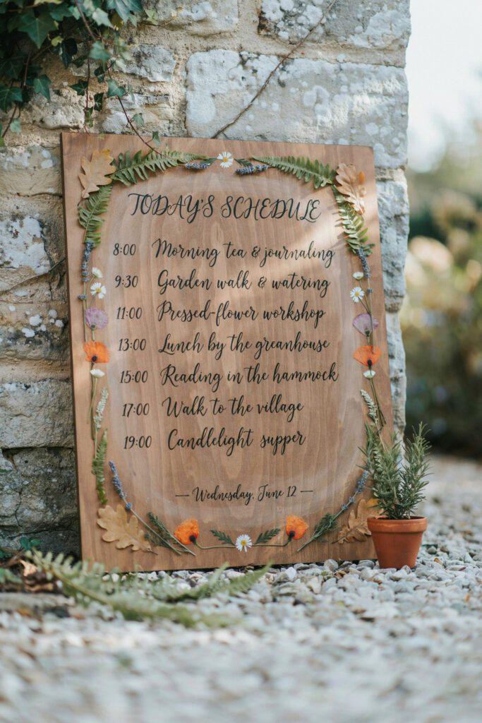  A large wooden board leaning against a garden wall with hand-lettered calligraphy listing the day's schedule, surrounded by a pressed botanical border of dried leaves, ferns, and small flowers arranged like an illustration frame