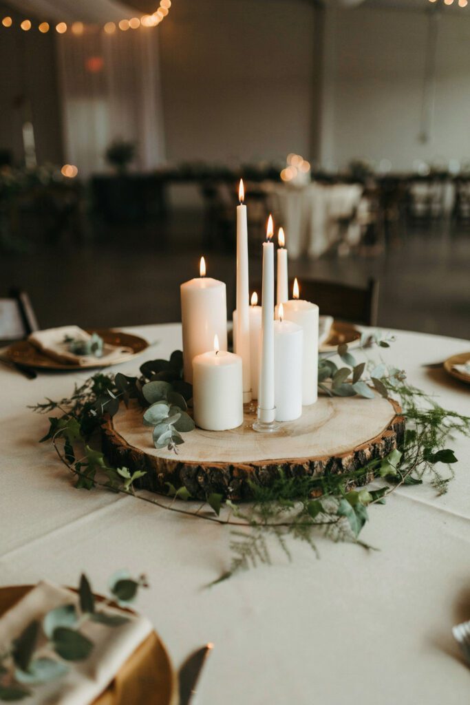 A reception table centerpiece on a thick wooden slice base, with a low arrangement of mixed greenery — eucalyptus, fern, and ivy — spreading outward from the base with a cluster of white candles in the center, no tall flowers