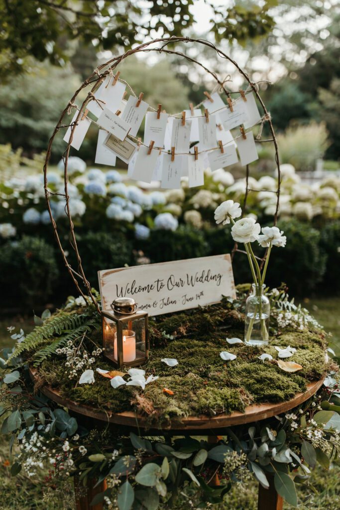 A round welcome table covered in moss and greenery, with a wooden "Welcome to Our Wedding" sign, a small lantern, a bud vase, and escort card display made from a wire vine frame with cards attached by tiny clips