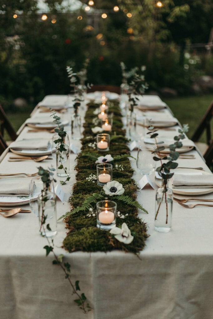 A long reception table with a continuous moss table runner down the center, interspersed with short glass votives holding floating candles, small clusters of fern fronds, and a few white anemones tucked in at intervals