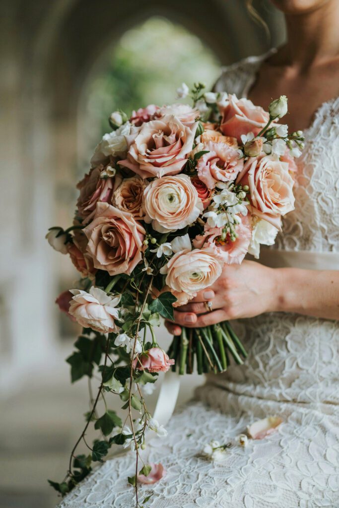 A lush, cascading bridal bouquet held against a white lace gown, featuring blush garden roses, pale peach ranunculus, and long trails of ivy and jasmine vine falling past the bride's hands