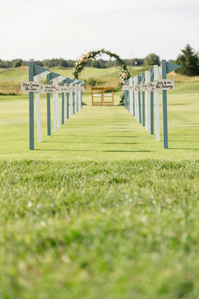 A grass aisle flanked by golf flagsticks in alternating white and sage green, each holding a small wooden hanging sign with directional or personal messages — "Here Comes the Sun," "Love Is on the Fairway," the couple's wedding date.