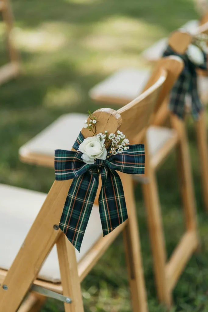 Close-up of ceremony chair backs tied with wide tartan plaid ribbon in navy and forest green — small sprig of white flowers tucked into the bow, sunlight on the course grass behind.