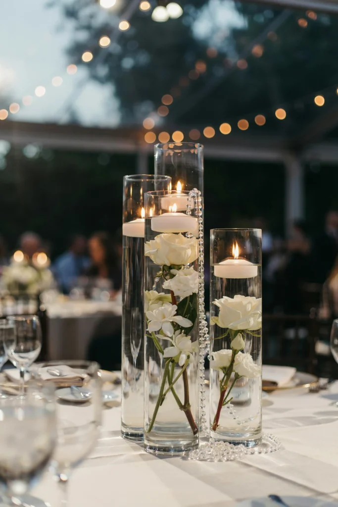 A reception centerpiece of tall cylindrical vases filled with water, floating ivory candles, and submerged white flowers — gardenias and roses — with crystal beads draped over the rim