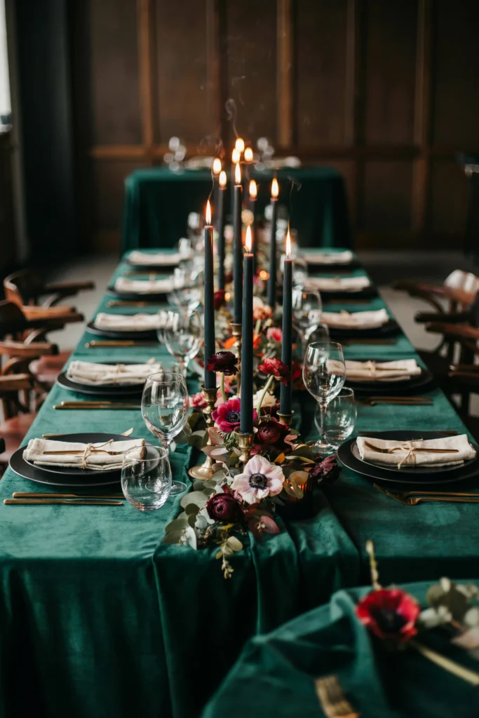 A moody dinner reception with emerald green velvet linens, tall black taper candles in brass candleholders, jewel-toned anemone and ranunculus centerpieces, and dark wooden chairs