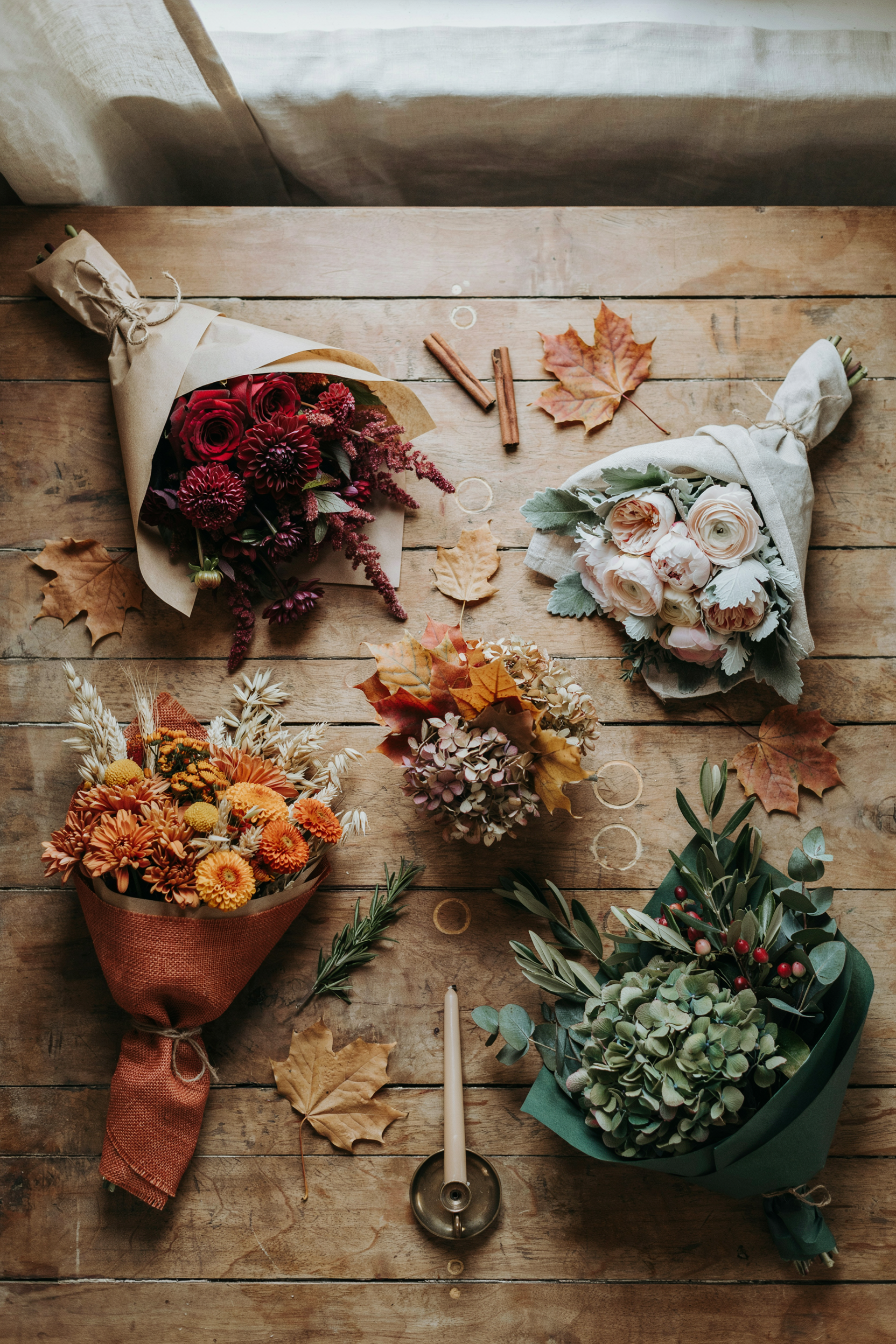 A curated flat lay or styled shot showing 4–6 diverse fall bouquets representing the full color range (deep burgundy, blush, burnt orange, forest green). Warm, natural window light. High-quality, Pinterest-worthy