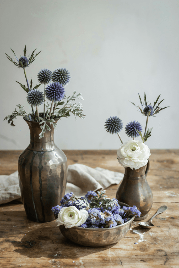 Eclectic arrangements in aged pewter vessels combining Scottish thistle, dusty blue globe thistles, dried blue statice, and silver artemisia with white ranunculus accents