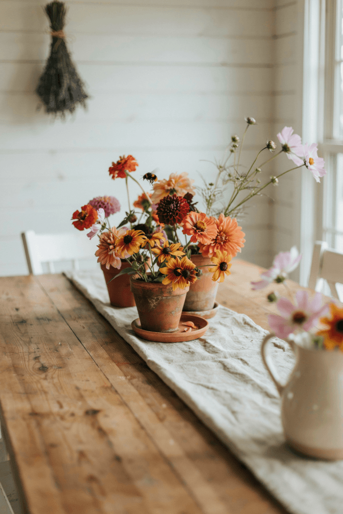 Casual, lush wildflower arrangements spilling out of terra cotta pots — zinnias, rudbeckia, dahlias, and cosmos in a riot of warm tones — on a table with a natural linen runner.