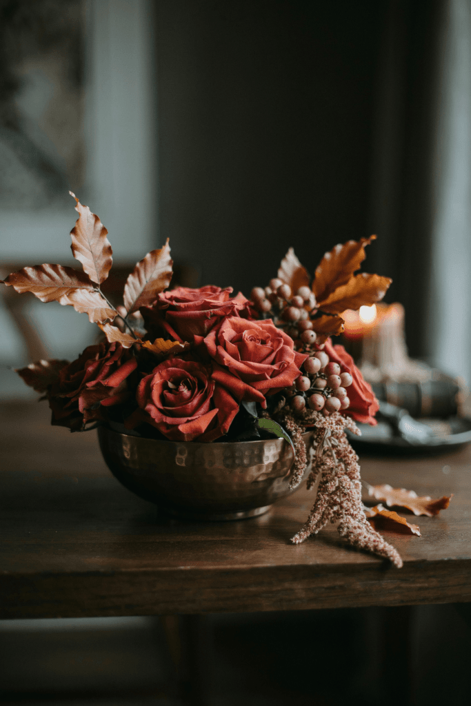 A large low arrangement in a hammered bronze compote, featuring oxblood roses, velvet amaranthus trailing over the edge, copper beech leaves, and small clusters of dusty mauve berries
