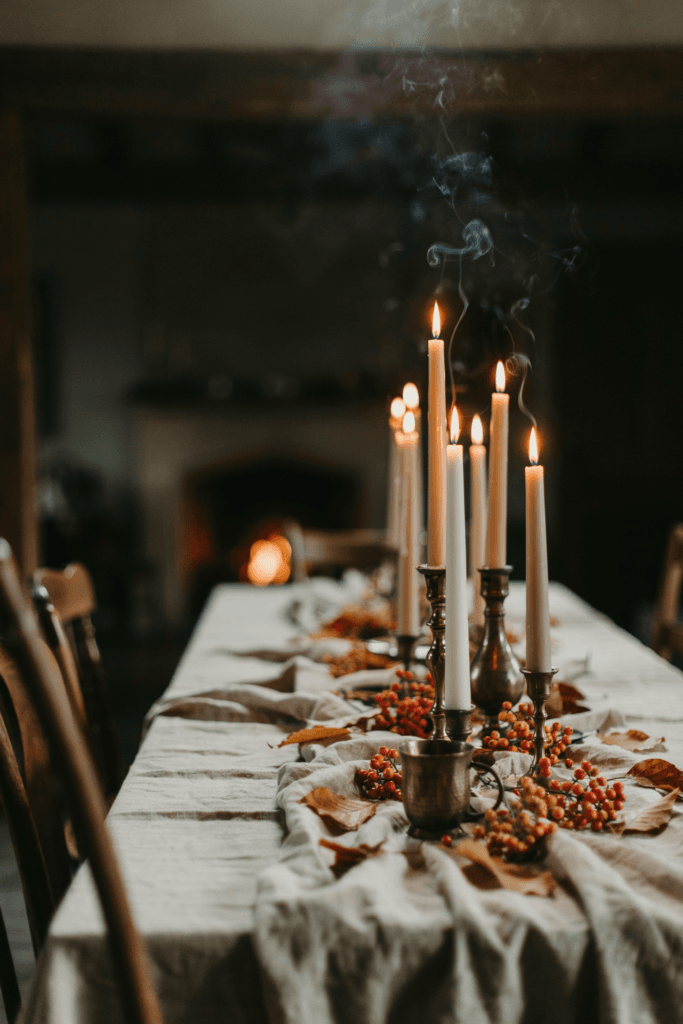 A tablescape dominated by clusters of tall taper candles in mismatched brass holders at varying heights, surrounded by loose hypericum berry stems and scattered autumn leaves in amber and gold