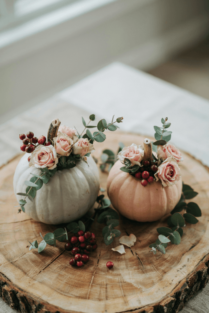 Hollow white and blush pumpkins used as vases, each holding a small arrangement of garden roses, eucalyptus, and small berries on a wood slice base