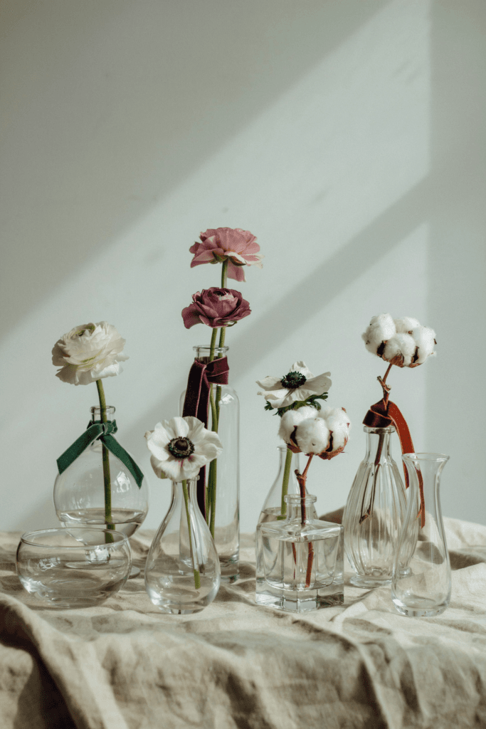  A cluster of 7–9 glass bud vases in varying heights and shapes, each wrapped with a single strip of deep velvet ribbon in plum, hunter green, or rust, holding single stems of ranunculus, anemones, and cotton bolls