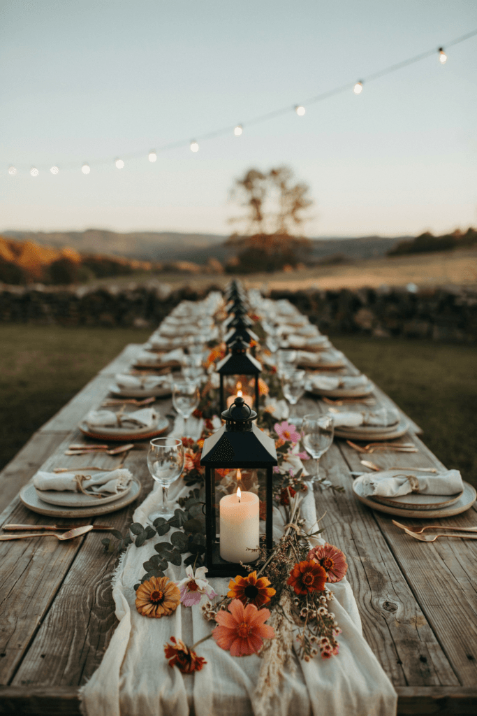 Black iron lanterns with pillar candles surrounded by loose wildflower arrangements in amber and terracotta tones — zinnias, rudbeckia, and cosmos — at a rustic outdoor fall reception