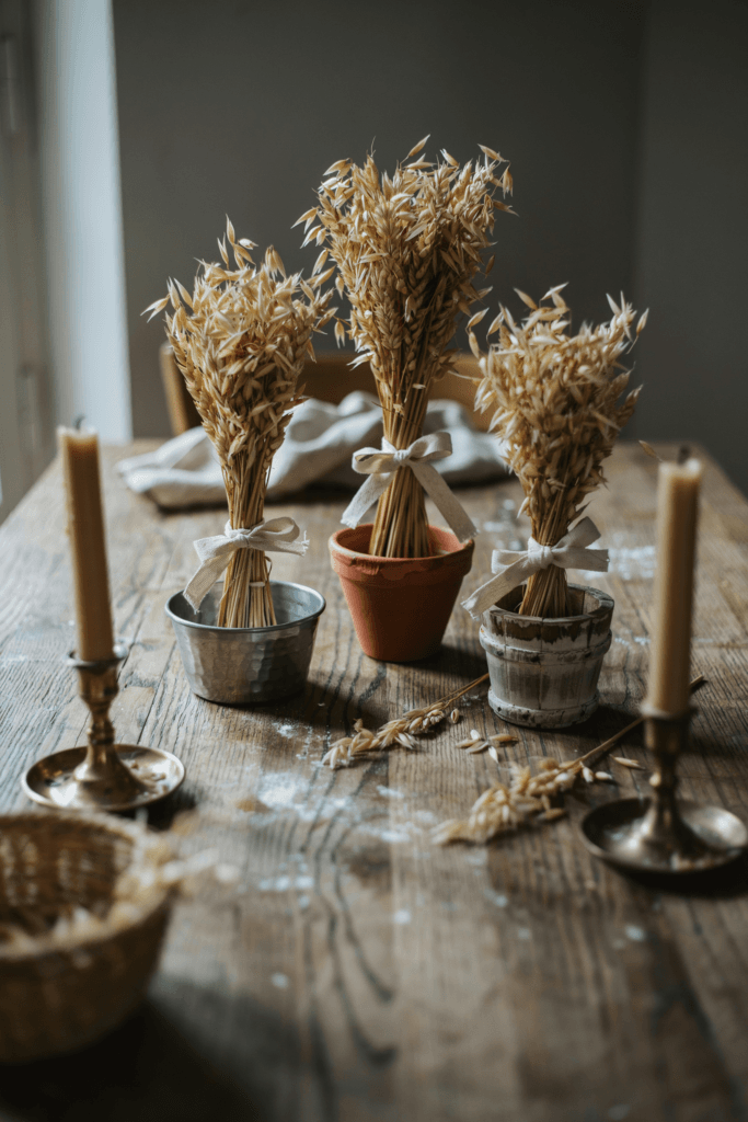Gathered sheaves of dried wheat tied with natural linen ribbon at the base, standing upright in shallow round vessels beside single candles in brass holders