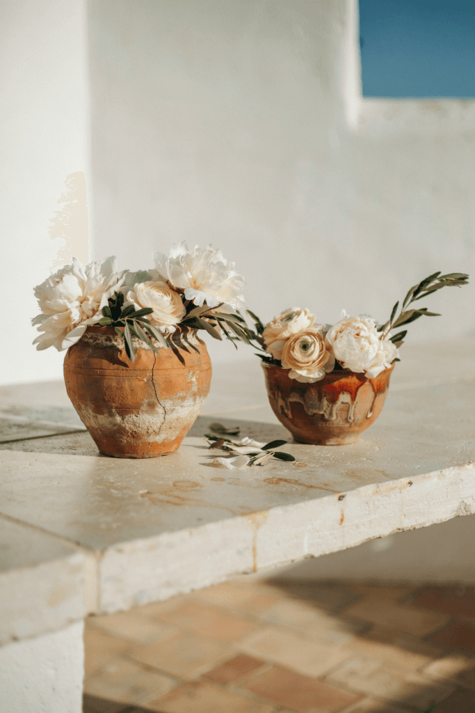  Low arrangements combining olive branches with full white peonies and cream ranunculus in aged terracotta vessels with a Mediterranean-meets-autumn feel