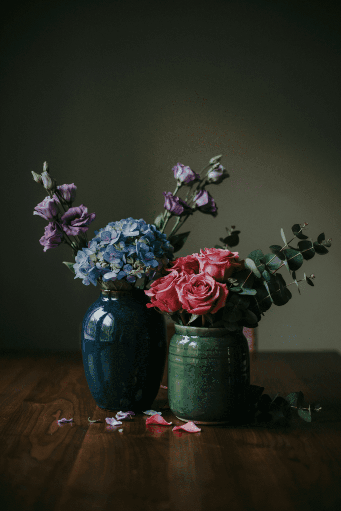 Low arrangements in deep navy and forest green glazed ceramic vases, filled with jewel-toned blooms — sapphire-blue hydrangea, amethyst lisianthus, ruby-toned roses, and deep hunter green foliage
