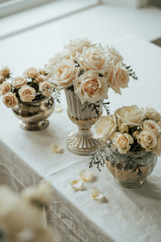 Ornate compote bowl vases overflowing with ivory garden roses, champagne spray roses, and wisps of silver-green dusty miller on a white linen tablecloth