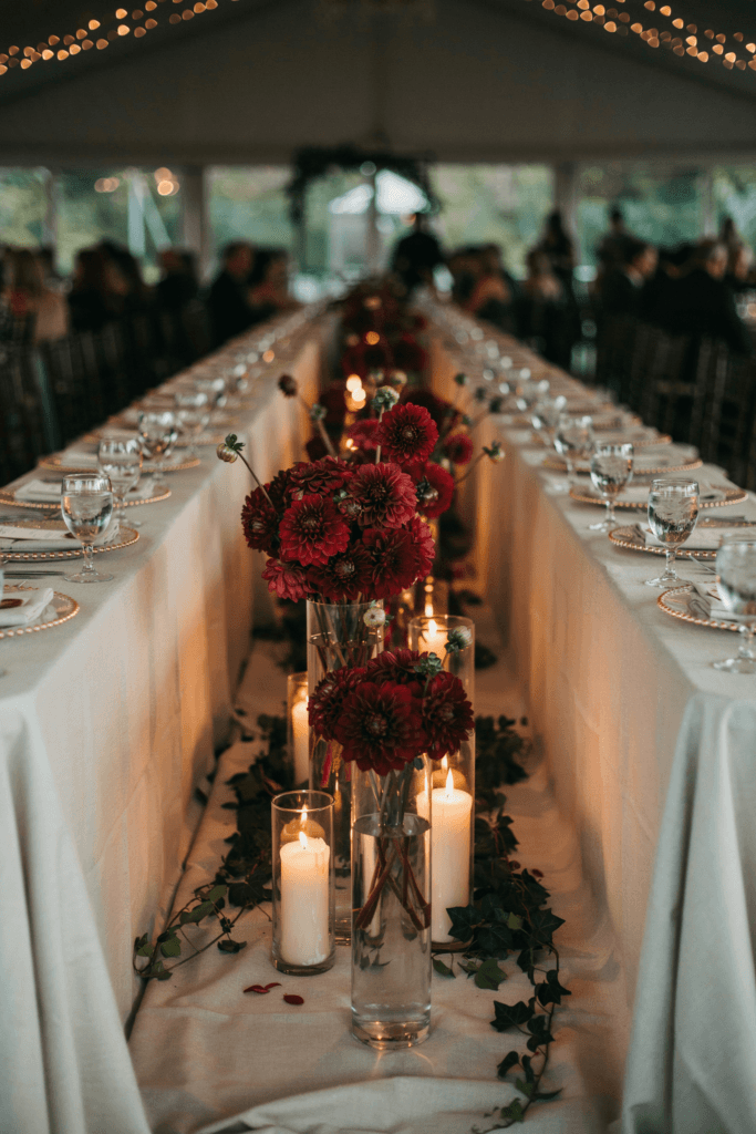 Tall glass cylinder vases filled with dense burgundy dahlia arrangements and trailing ivy at a candlelit reception table