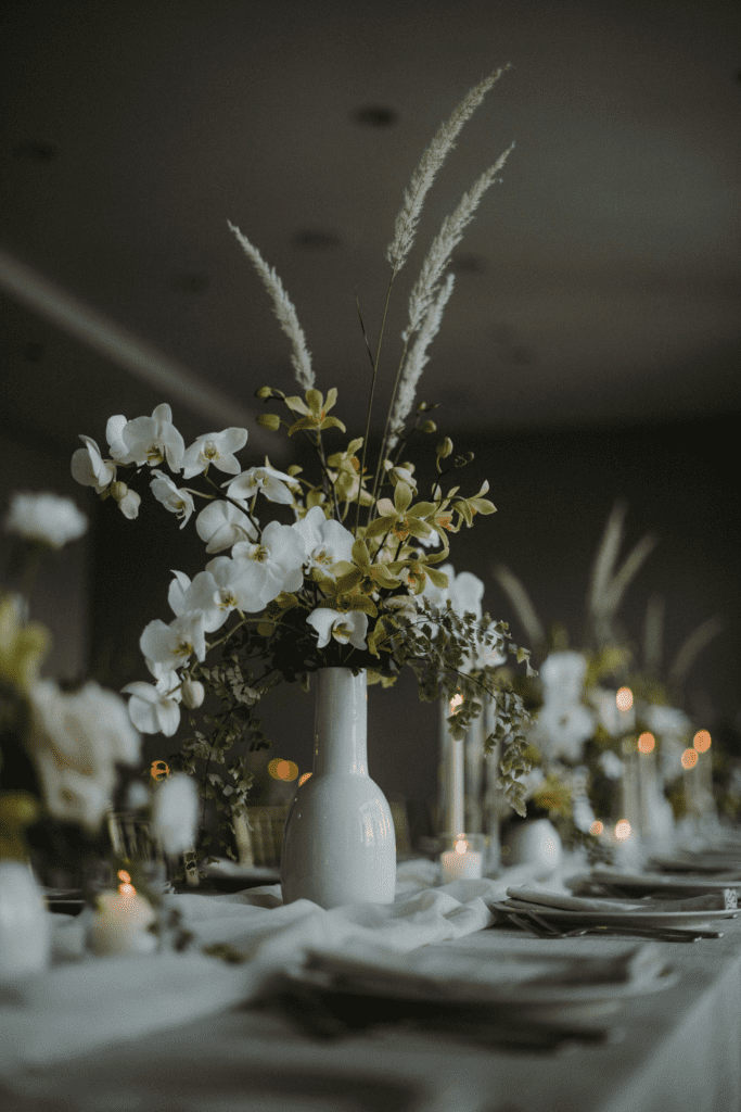 Tall, dramatic centerpieces in slim white lacquered vessels with cascading white and chartreuse orchid stems, trailing maidenhair fern, and single stems of bear grass creating an arching silhouette