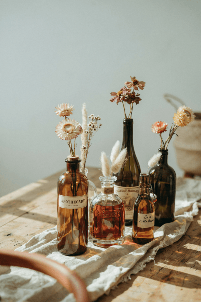 Eclectic vintage bottle collections — amber, cognac, and dark green glass — each holding 1–3 stems of dried strawflowers, pampas, or single cosmos, grouped in loose clusters on a table runner