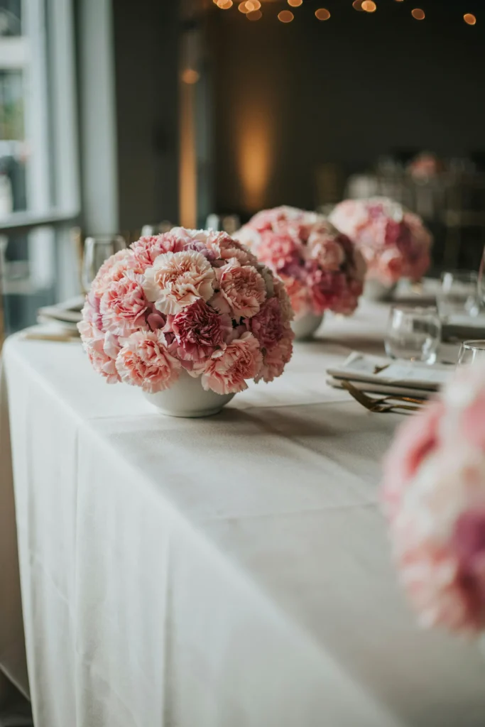 Lush, full centerpieces made entirely of tightly packed pink carnations in a gradient from pale blush to deep rose, in short white ceramic vessels