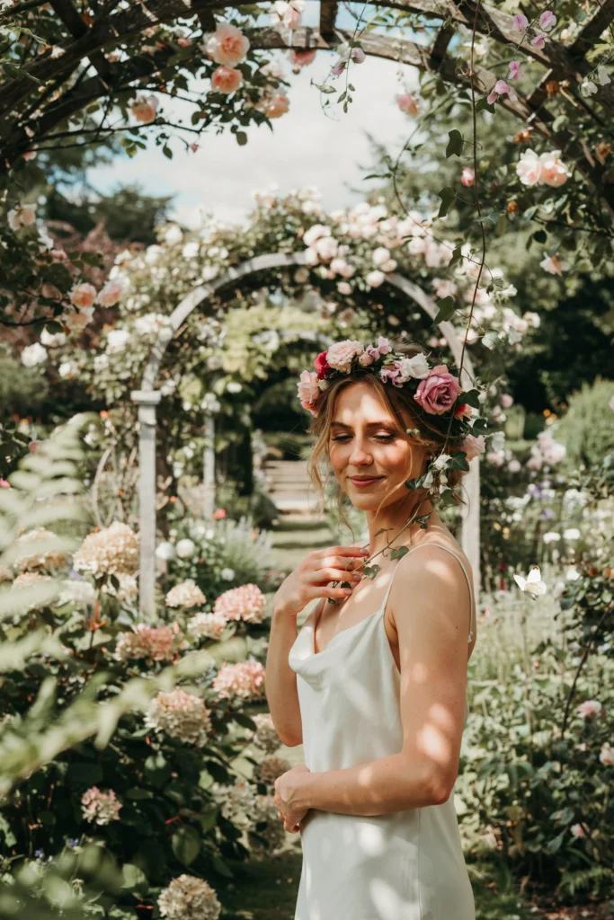 A relaxed bridal portrait with the bride wearing a loose flower crown of mixed pink garden roses, sweet peas, and trailing greenery against a lush outdoor backdrop