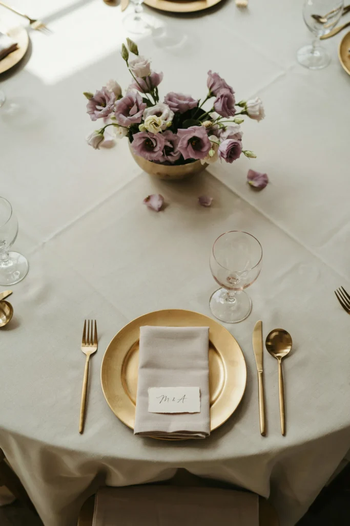 A beautifully set round table with a champagne linen, mauve cloth napkin, a blush goblet, and a low centerpiece of mauve lisianthus and ivory garden roses in a low gold bowl