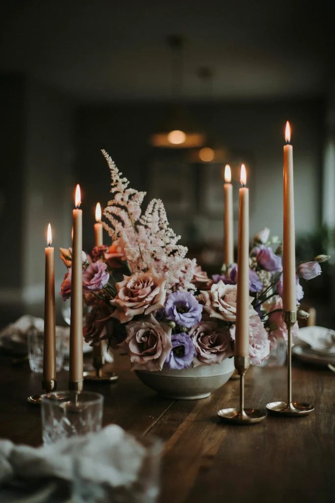 A low, lush centerpiece of mauve amnesia roses, astilbe, and lisianthus surrounded by gold taper candles in slender brass candleholders