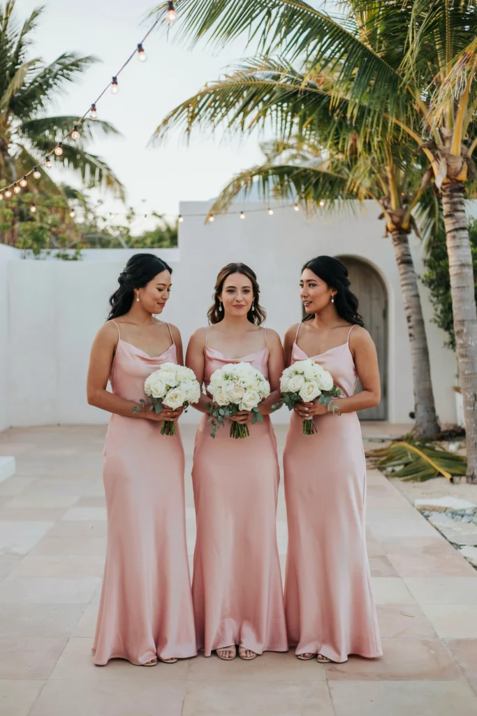 Three bridesmaids in floor-length hot pink satin dresses holding tight, round all-white bouquets of garden roses against a palm-lined outdoor venue