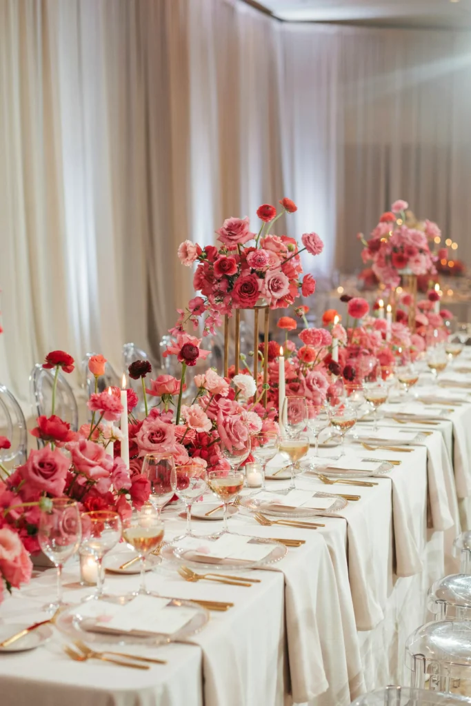 A banquet table overflowing with hot pink florals in varying heights, white pillar candles, white linens, gold flatware, and a champagne coupe at each setting