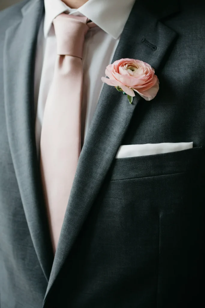A close-up of a groom's jacket lapel with a blush silk tie, a white pocket square, and a small pink ranunculus boutonnière — subtle and cohesive without competing with the bride