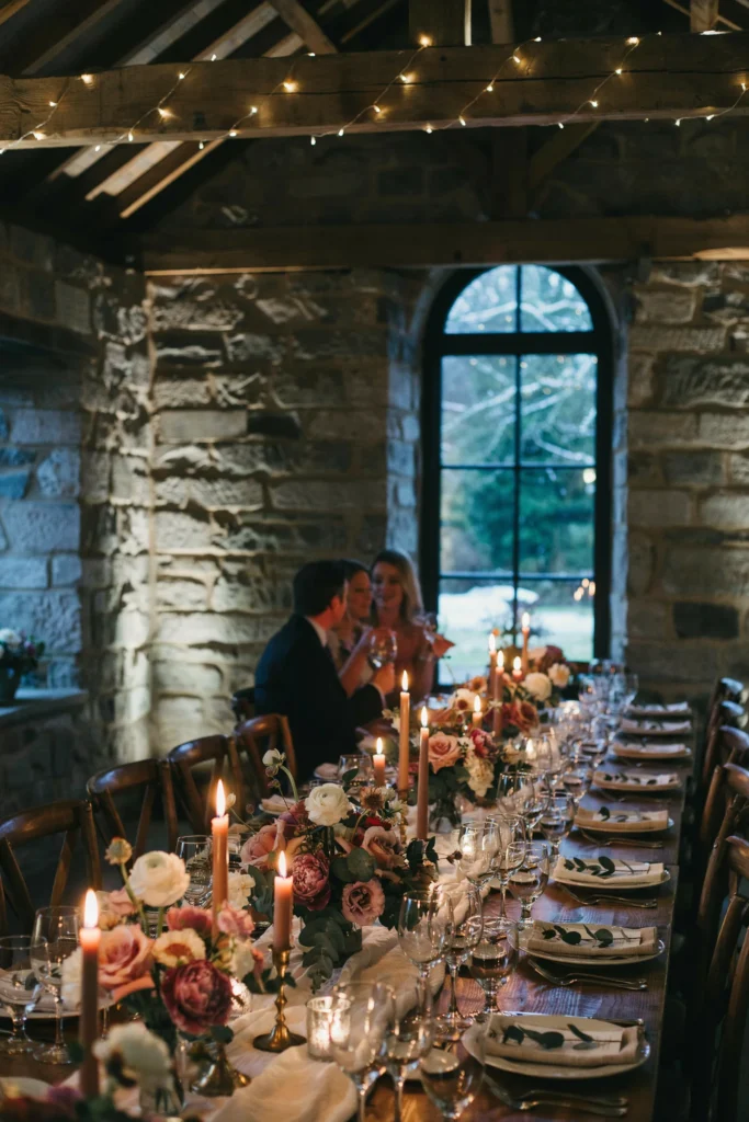 An intimate winter wedding reception in a stone-walled venue, tables lit entirely by candlelight, with mauve and white florals glowing warmly against the candlelit walls
