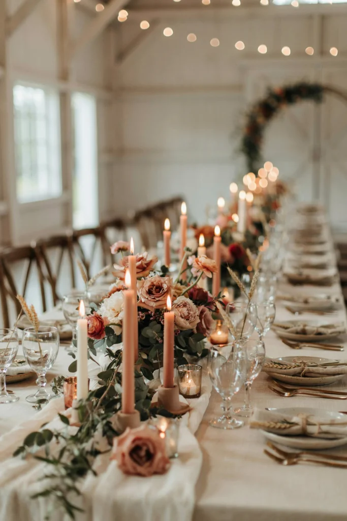 A fall wedding tablescape with dusty rose florals, terracotta-toned candles, deep green foliage, and warm amber lighting creating a rich and intimate atmosphere
