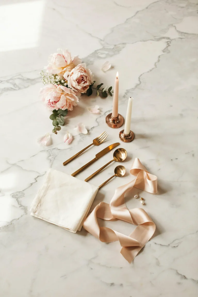 A dreamy styled wedding flat lay featuring blush peonies, rose gold candle holders, gold flatware, and a blush silk ribbon on a marble surface.