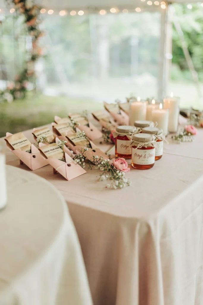 A display table of small wedding favors — seed packets in pink envelopes, small jars of pink honey, and mini candles — arranged on a blush linen with a floral sprig at each one