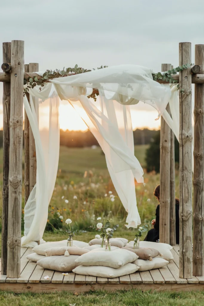 Outdoor ceremony or lounge area with long lengths of white chiffon fabric draped between wooden posts, billowing gently in a breeze, with minimal floral accents and white cushioned seating below